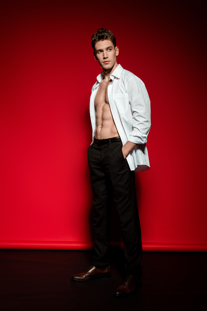sexy joven elegante hombre en camisa desabotonada con torso desnudo muscular y las manos en bolsillos sobre fondo rojo
 - Foto, imagen