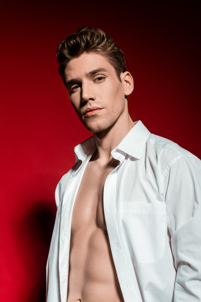 sexy young elegant man in unbuttoned shirt with muscular bare torso on red background - Photo, Image