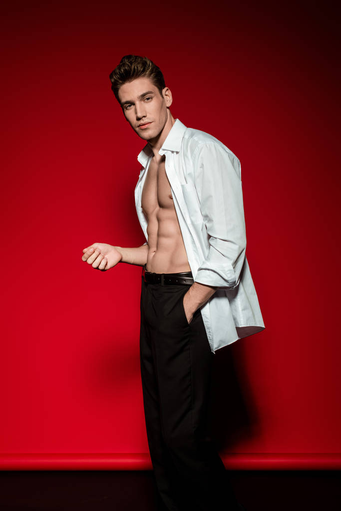 sexy jovem elegante homem em camisa desabotoada com muscular tronco nu posando com a mão no bolso no fundo vermelho
 - Foto, Imagem