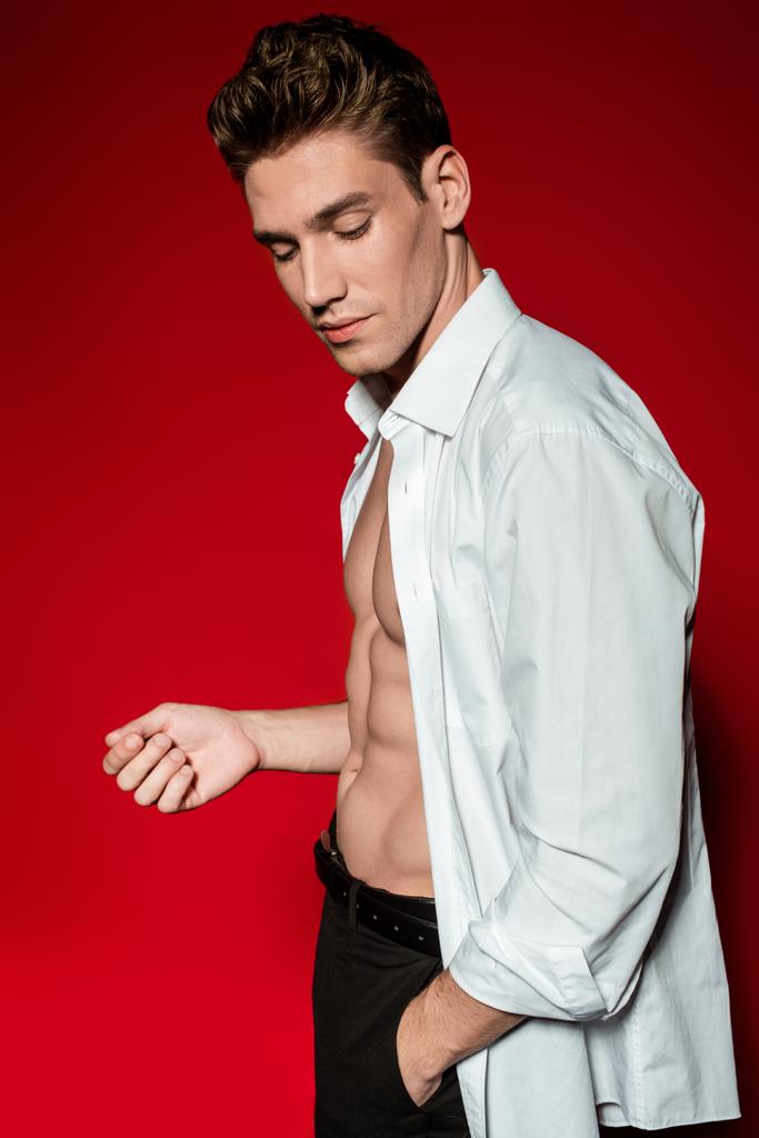 baixo ângulo vista de sexy jovem elegante homem em camisa desabotoada com muscular tronco nu posando com a mão no bolso no fundo vermelho
 - Foto, Imagem
