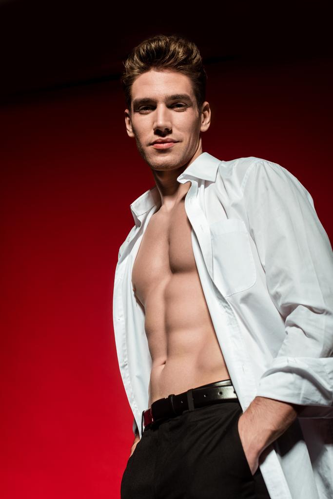 άποψη χαμηλή γωνία του σέξι νεαρός κομψό άνδρα σε ξεκούμπωτο πουκάμισο με μυώδη γυμνό κορμό θέτουν με τα χέρια σε τσέπες στο κόκκινο φόντο - Φωτογραφία, εικόνα