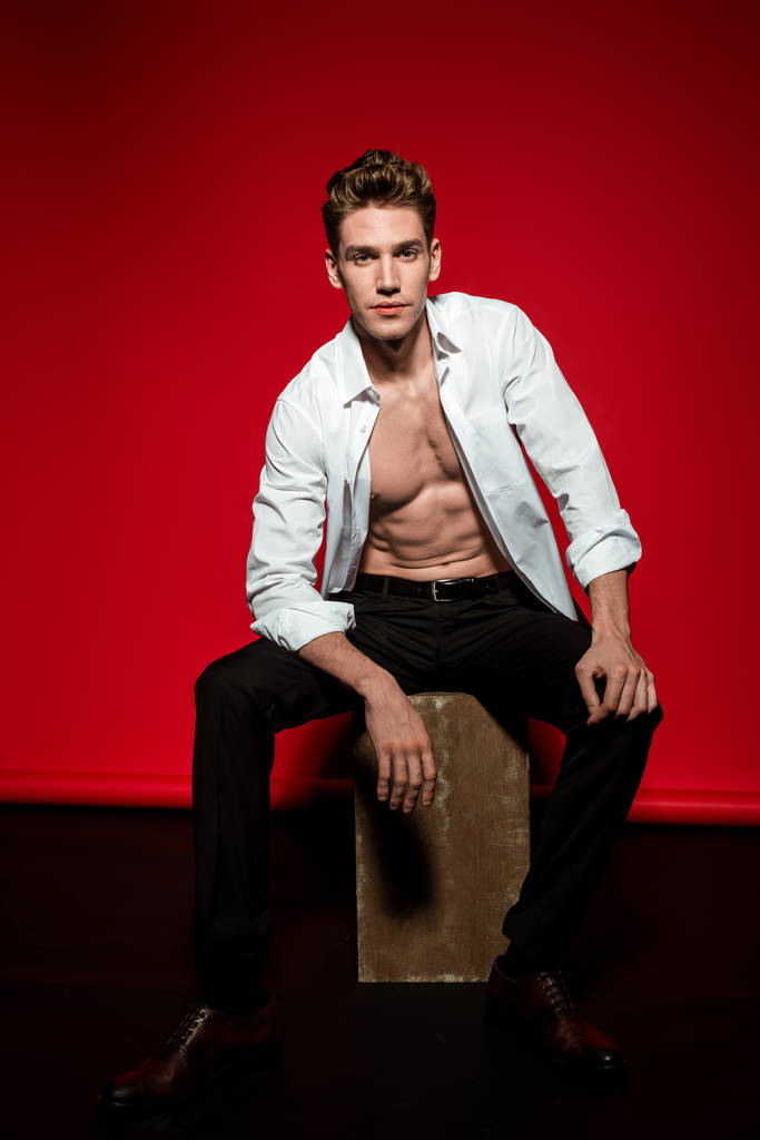 sexy junger eleganter Mann im aufgeknöpften Hemd mit muskulösem nacktem Oberkörper posiert auf Schachtel auf rotem Hintergrund - Foto, Bild