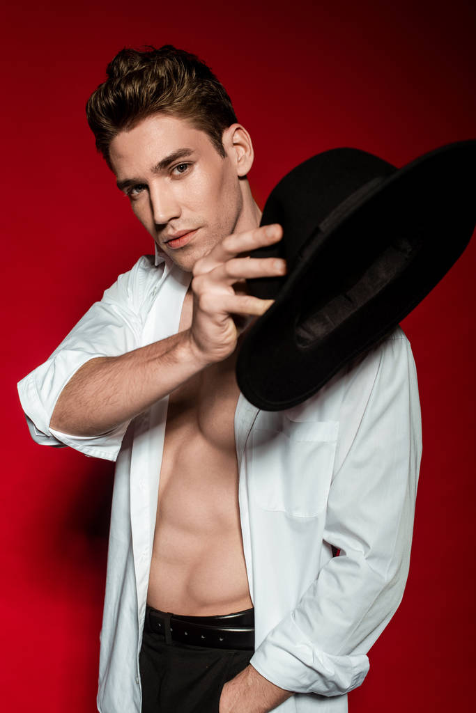 sexy young elegant man in unbuttoned shirt with muscular bare torso holding hat on red background - Photo, Image