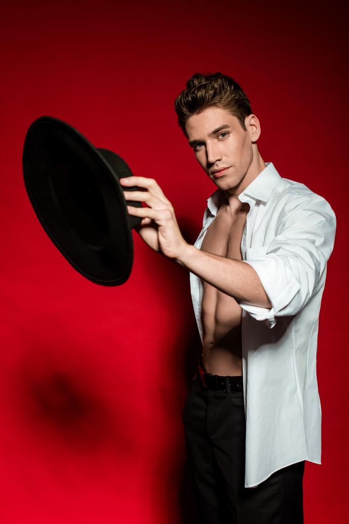 セクシーな若いですエレガントな男でunbuttonシャツとともに筋肉裸胴ホールディング帽子上の赤の背景 - 写真・画像