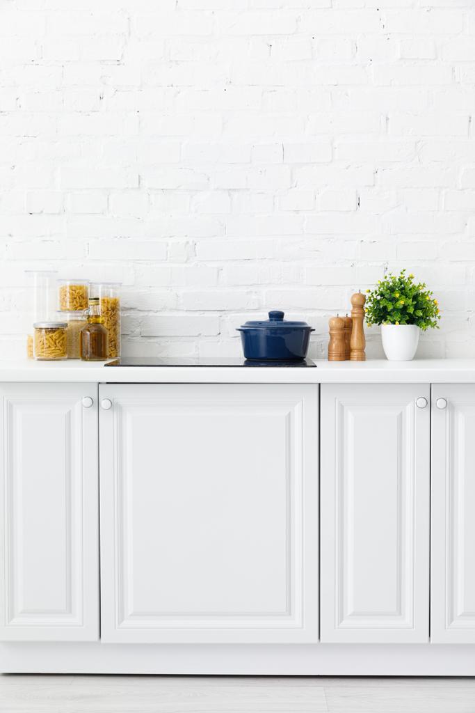 μοντέρνο λευκό κουζίνα εσωτερικό με κατσαρόλα σε ηλεκτρική επαγωγική βάση εστιών κοντά στη διακόσμηση και δοχεία τροφίμων κοντά στον τοίχο από τούβλα - Φωτογραφία, εικόνα