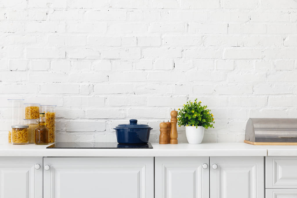 interior de cozinha branca moderna com panela em fogão de indução elétrica perto de recipientes de plantas e alimentos perto da parede de tijolo
 - Foto, Imagem