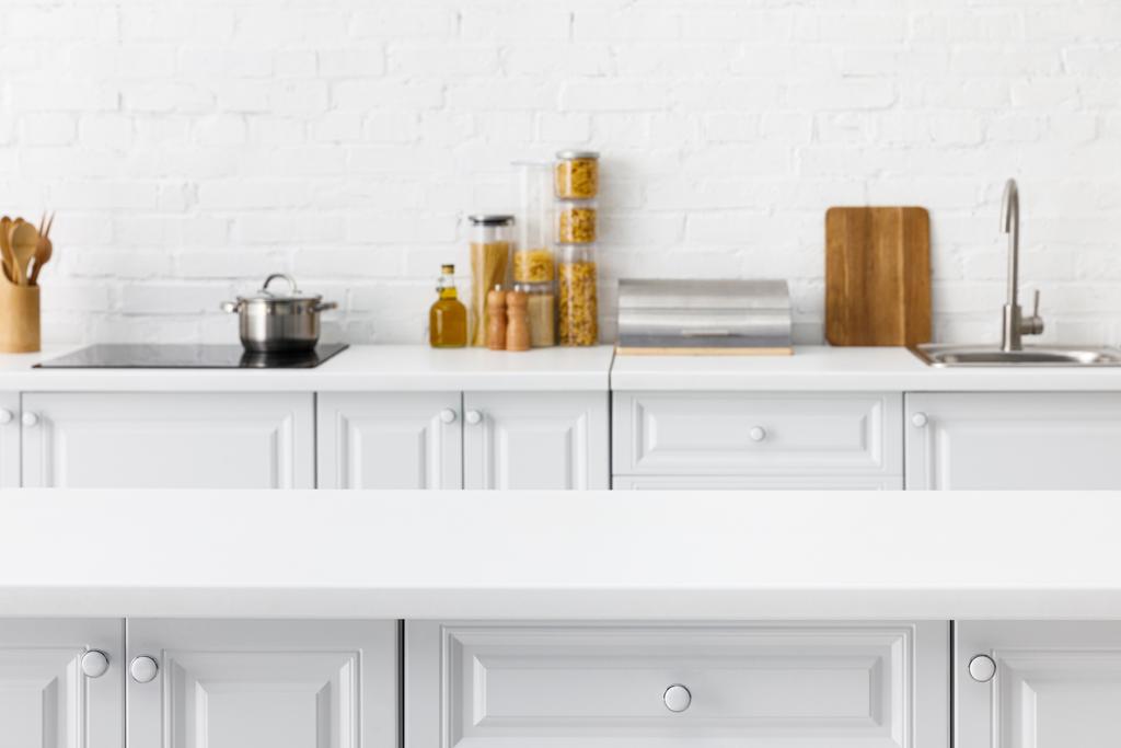 selective focus of empty tabletop and minimalistic modern white kitchen interior with kitchenware and food near brick wall on background - Photo, Image