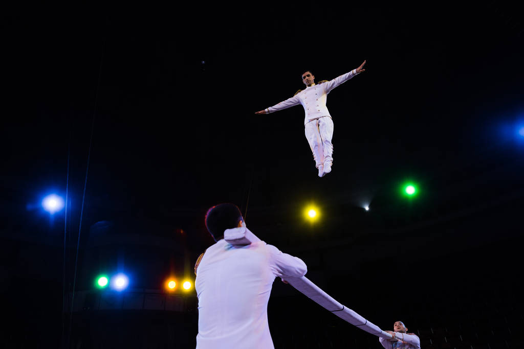 niski kąt widzenia akrobata powietrza z wyciągniętymi rękami skaczących w pobliżu bieguna na arenie cyrku  - Zdjęcie, obraz