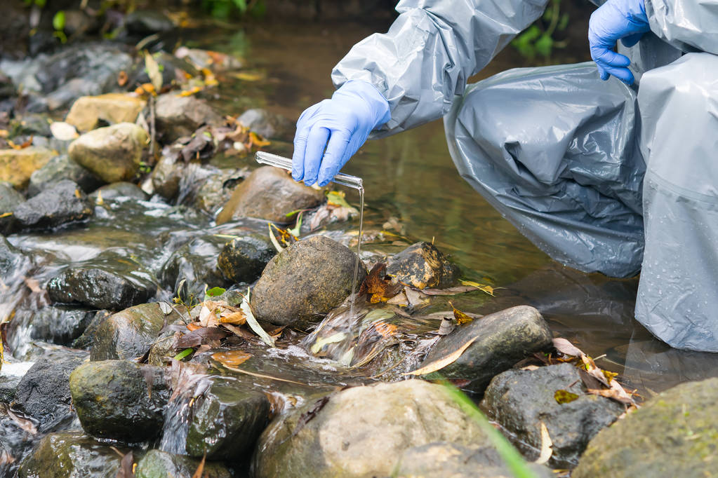 ein Mann in einem Schutzanzug steht in einem Fluss und schüttet Wasser aus einem Reagenzglas hinein, um den Grad der Verschmutzung zu analysieren - Foto, Bild