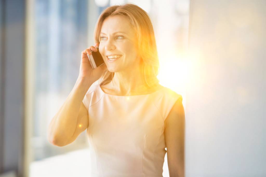 Mujeres de negocios sonriendo mientras hablaban por teléfono inteligente en el cargo con lentes amarillas en segundo plano. - Foto, Imagen