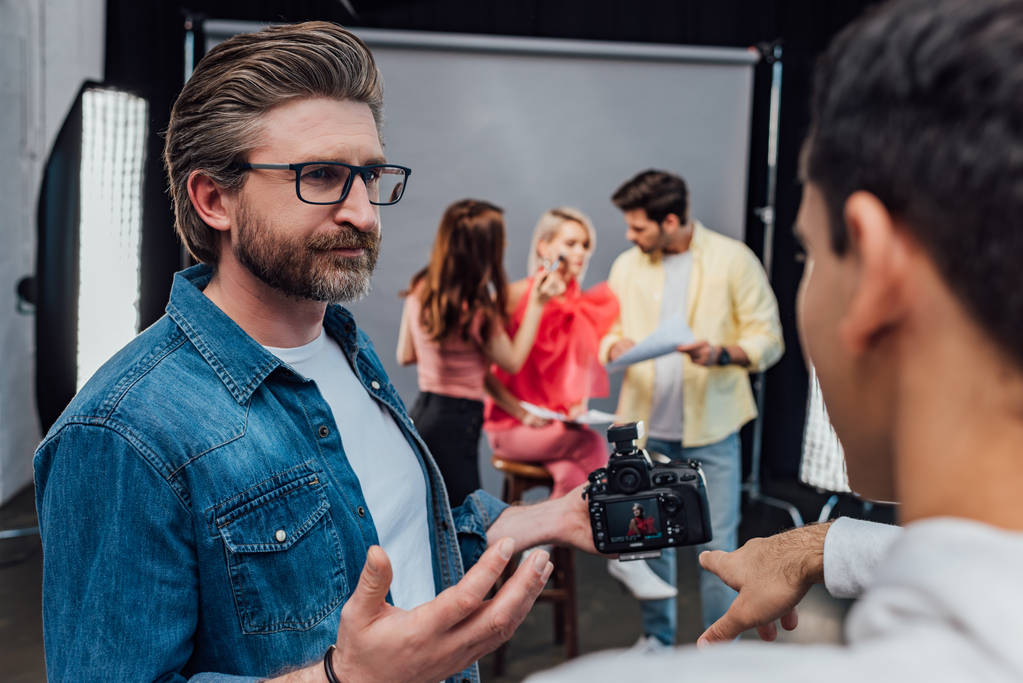 selective focus of bearded art director in glasses looking at assistant pointing with finger at digital camera  - Photo, Image