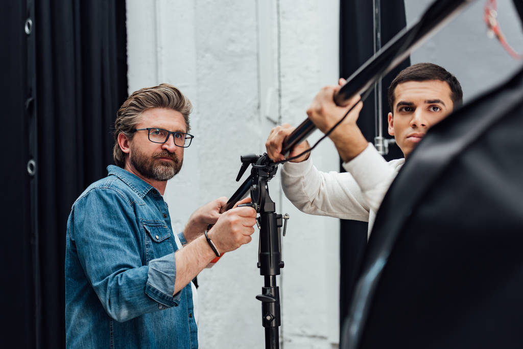 selektivní zaměření pohledných mužů držících lehký stojan ve fotostudiu  - Fotografie, Obrázek
