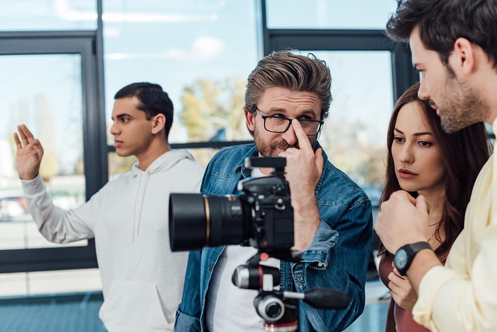 επιλεκτική εστίαση του καλλιτεχνικού διευθυντή αγγίζοντας γυαλιά και κοιτάζοντας βοηθός κοντά ψηφιακή φωτογραφική μηχανή  - Φωτογραφία, εικόνα
