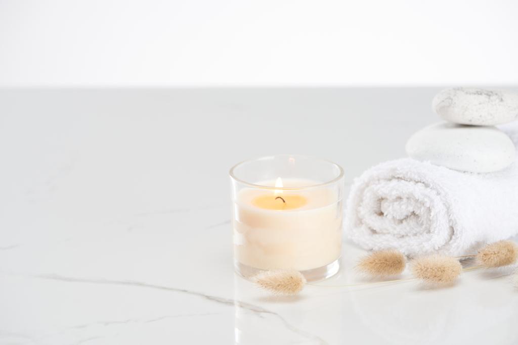 flauschiges Hasenschwanzgras in der Nähe brennender weißer Kerze im Glas und gerolltem Handtuch mit Steinen auf marmorweißer Oberfläche - Foto, Bild