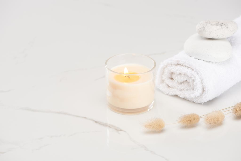 пушистая трава хвоста кролика возле горящей белой свечи в стекле и свернутое полотенце с камнями на мраморной белой поверхности
 - Фото, изображение