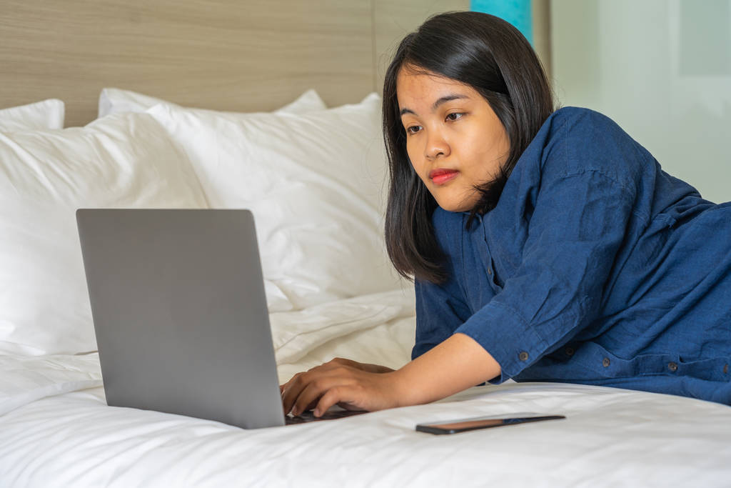 Fiatal nő fekszik az ágyon laptoppal mobiltelefon mellett - Fotó, kép