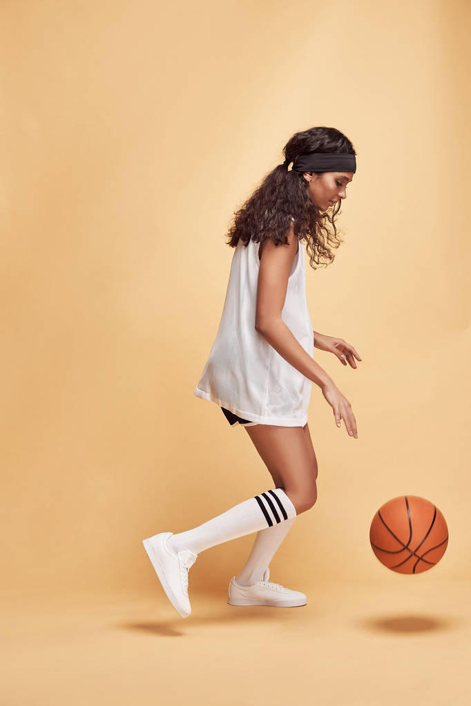 piękne ciemnoskóry dziewczyna stoi w pełnym wysokości na żółtym tle w białej koszuli sportowej, czarne szorty, białe wysokie skarpetki i tenisówki bawiące się piłkę do koszykówki - Zdjęcie, obraz
