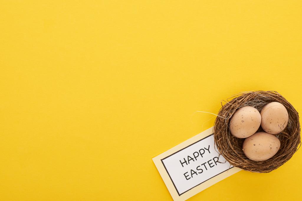 ylhäältä tervehdyskortti onnellisella pääsiäisellä kirjoituksella lähellä kananmunia pesässä keltaisella värikkäällä taustalla
 - Valokuva, kuva
