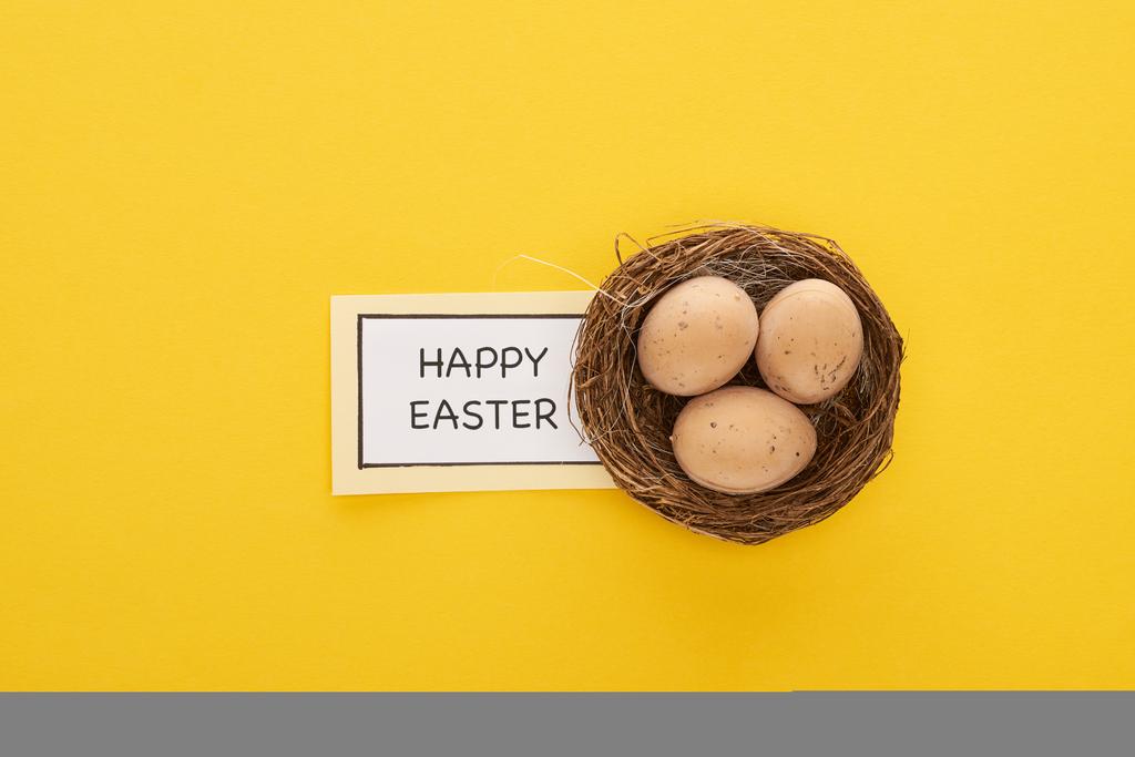 вид поздравительной открытки с радостными пасхальными надписями возле куриных яиц в гнезде на желтом красочном фоне
 - Фото, изображение