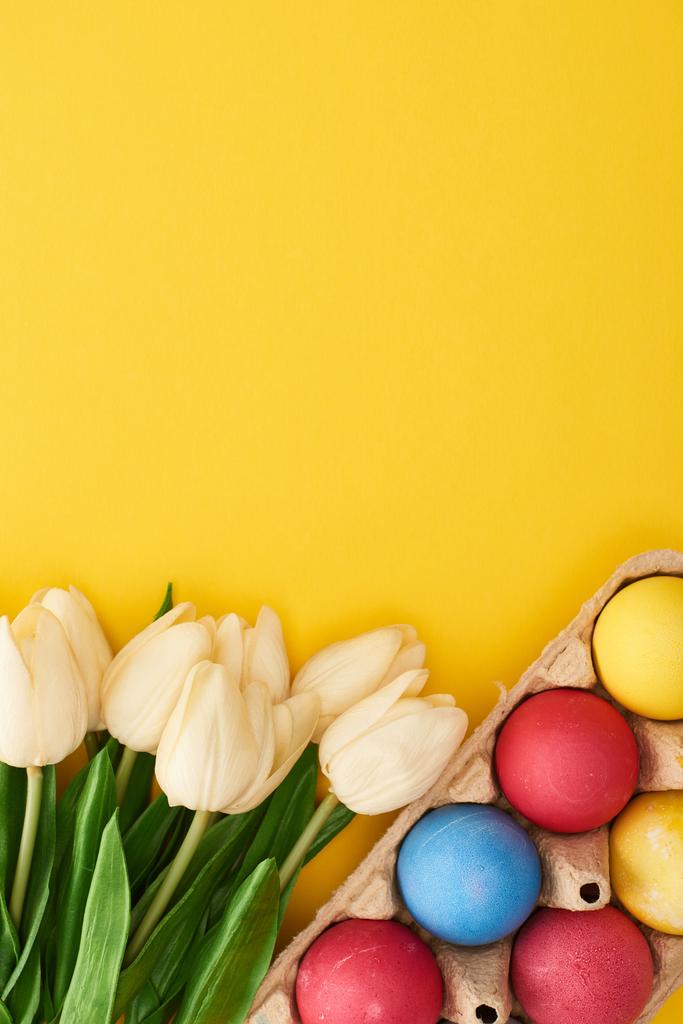 felső nézet tulipánok és színes festett húsvéti tojás kartondobozban színes sárga háttér - Fotó, kép