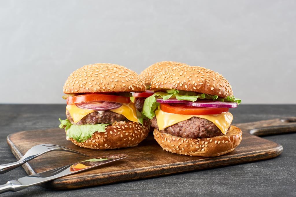 deliciosos cheeseburgers de carne fresca em tábua de madeira perto de talheres
 - Foto, Imagem