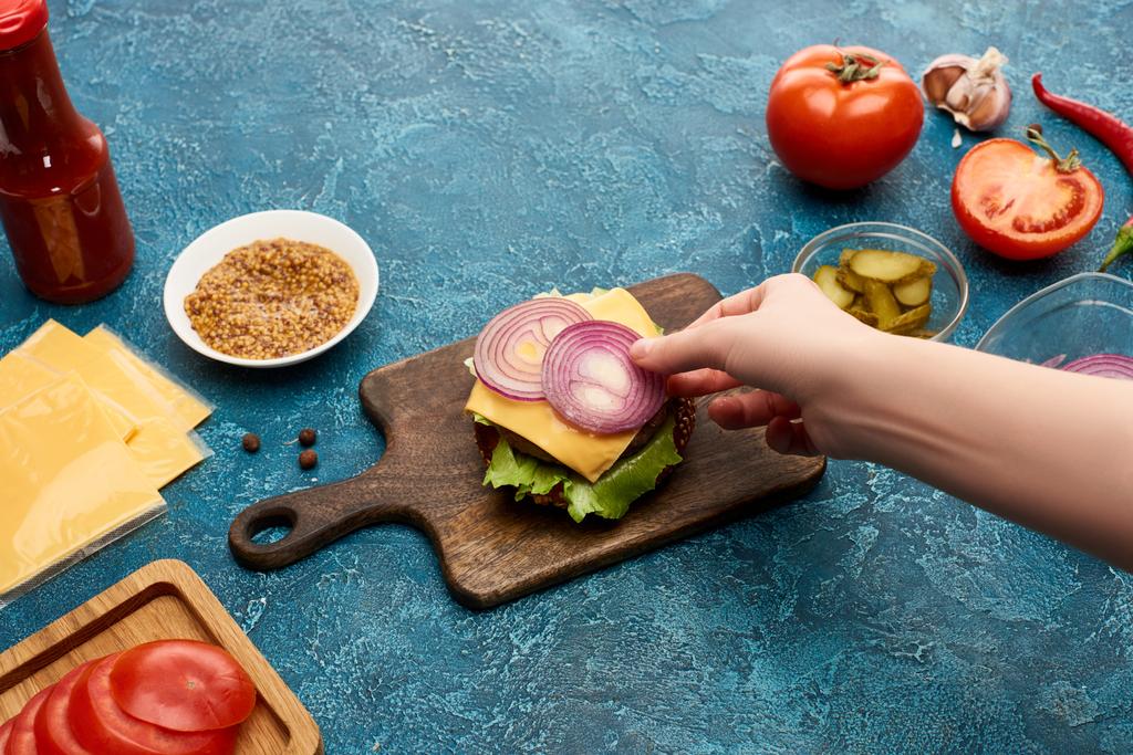 Mavi desenli yüzeyde taze hamburgerin üzerine soğan koyan kadının kırpılmış görüntüsü. - Fotoğraf, Görsel