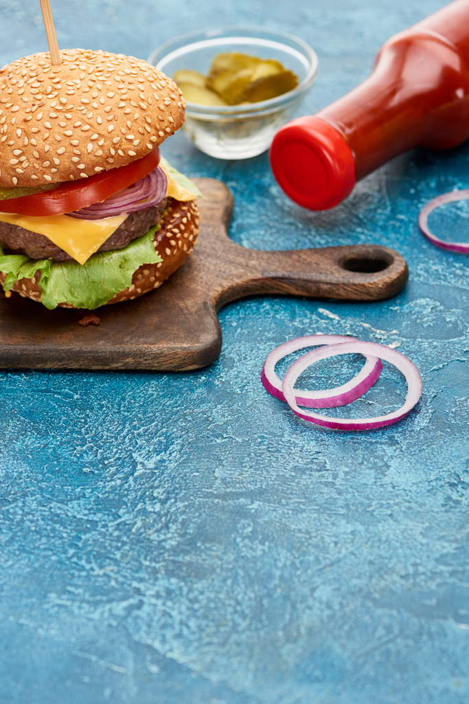 Turşu yakınındaki ahşap tahtada leziz çizburgerin ve mavi desenli yüzeyde ketçapın seçici odağı. - Fotoğraf, Görsel