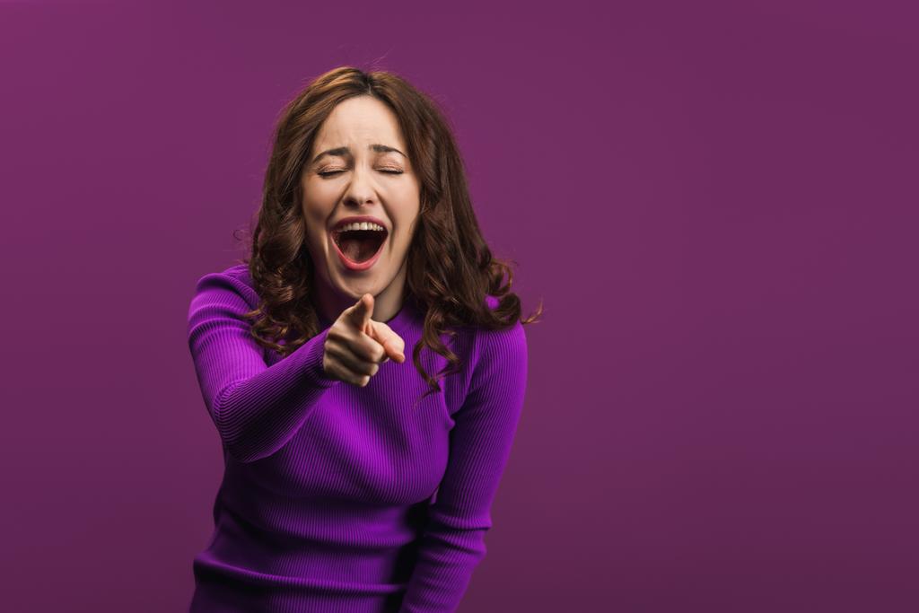 wesoła kobieta śmiejąca się z zamkniętych oczu i wskazująca palcem na kamerę na fioletowym tle - Zdjęcie, obraz