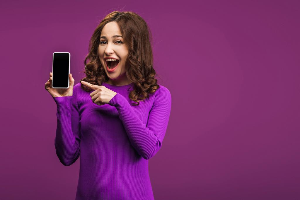 lachende Frau zeigt mit dem Finger auf Smartphone mit leerem Bildschirm auf lila Hintergrund - Foto, Bild