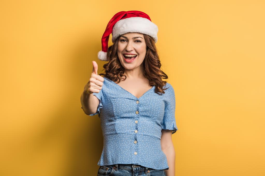 cheerful girl in santa hat showing thumb up on yellow background - Photo, Image