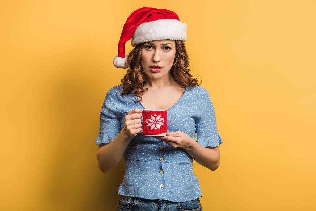 unzufriedenes Mädchen mit Weihnachtsmannmütze mit wärmendem Getränk auf gelbem Hintergrund - Foto, Bild