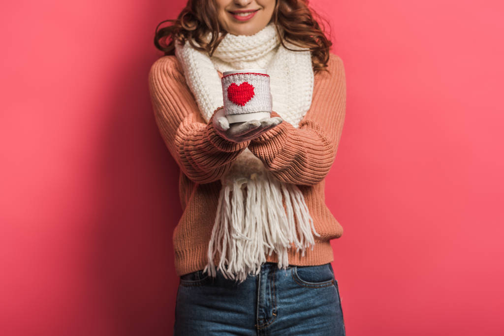 bijgesneden weergave van lachend meisje in warme sjaal holding cup met hart symbool op roze achtergrond - Foto, afbeelding