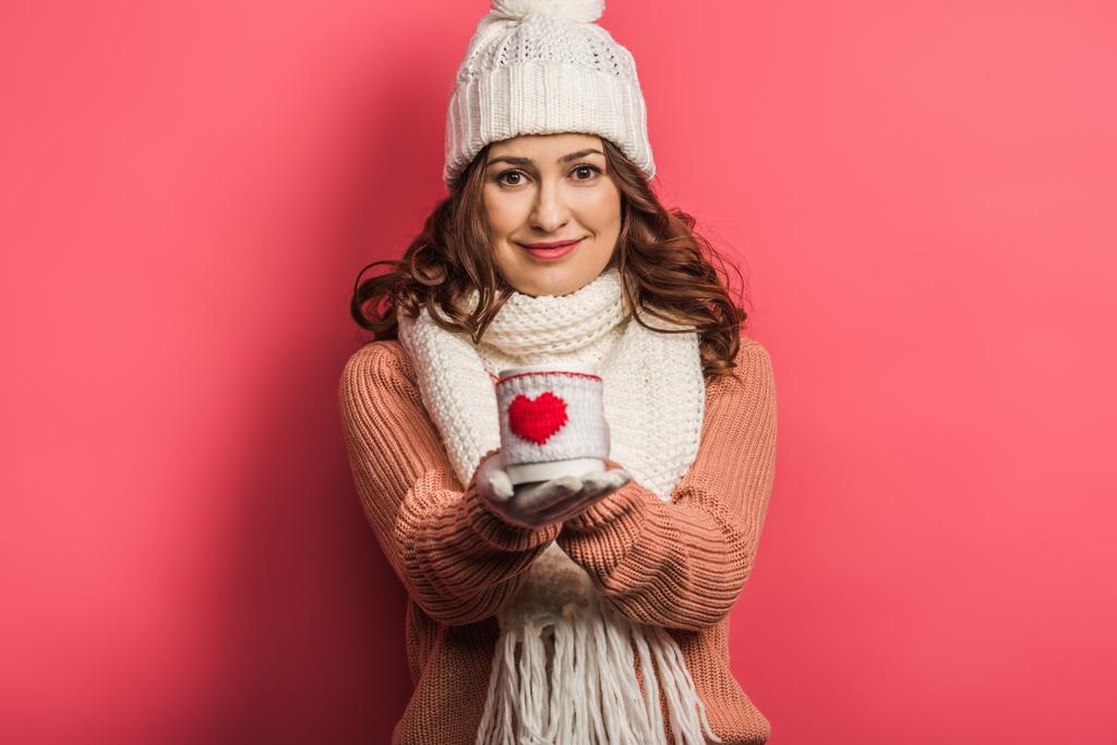 boldog lány meleg kalap és sál tartó csésze szív szimbólum rózsaszín háttér - Fotó, kép