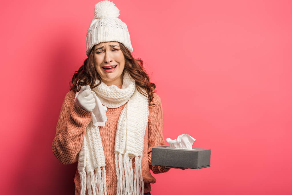 sad girl in warm hat and scarf crying while holding paper napkins on pink background - Photo, Image