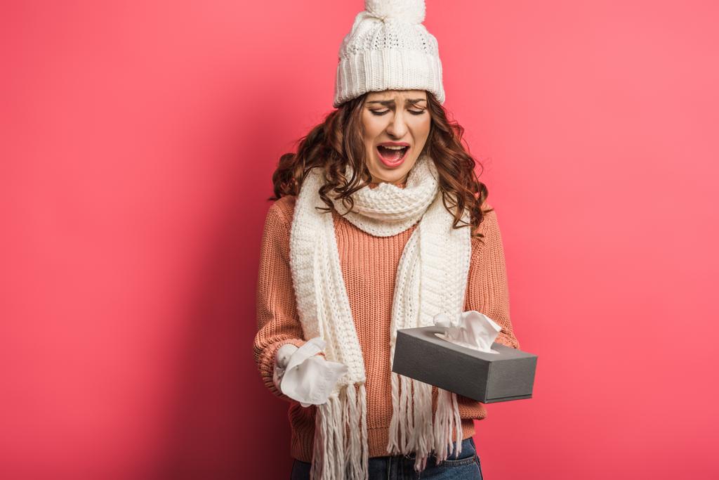 upset girl in warm hat and scarf crying while holding paper napkins on pink background - Photo, Image