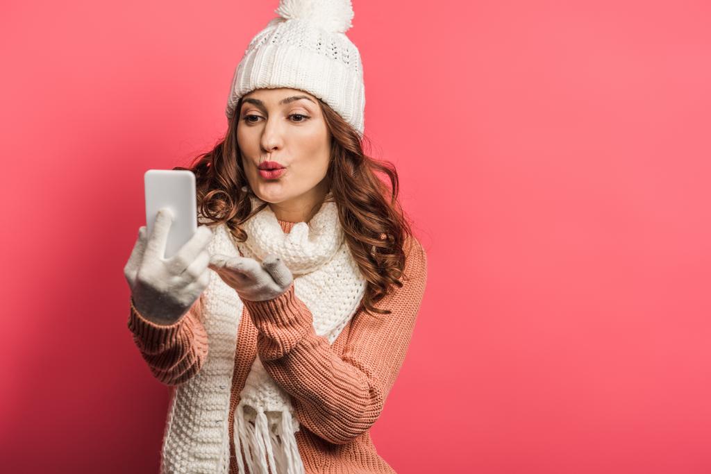 fille positive en chapeau chaud et écharpe envoyer baiser d'air pendant l'appel vidéo sur smartphone sur fond rose
 - Photo, image