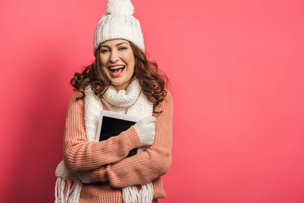cheerful girl in warm hat and scarf holding digital tablet on pink background - Photo, Image