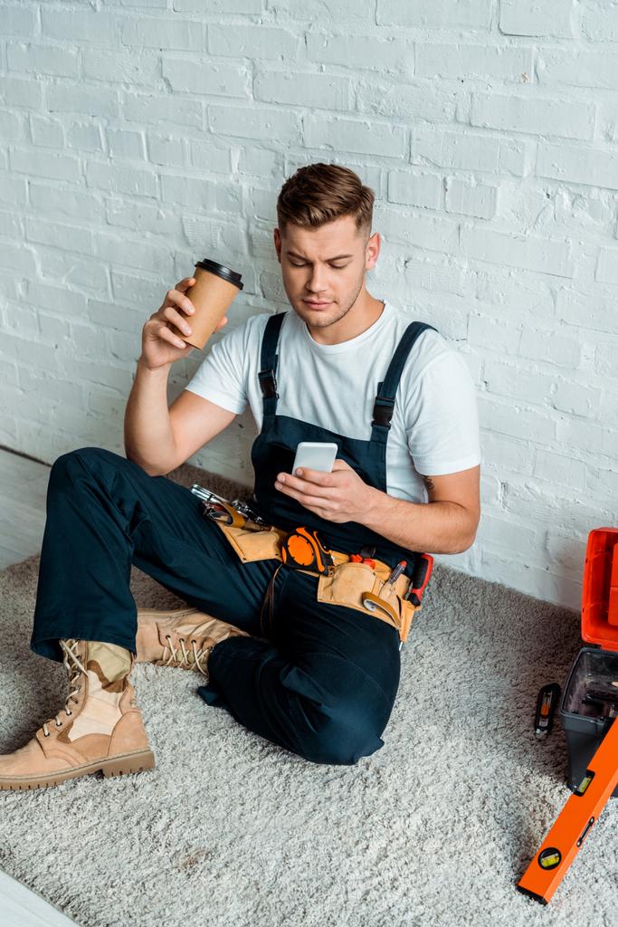 όμορφος εγκαταστάτης που χρησιμοποιεί smartphone ενώ κρατά χάρτινο κύπελλο  - Φωτογραφία, εικόνα