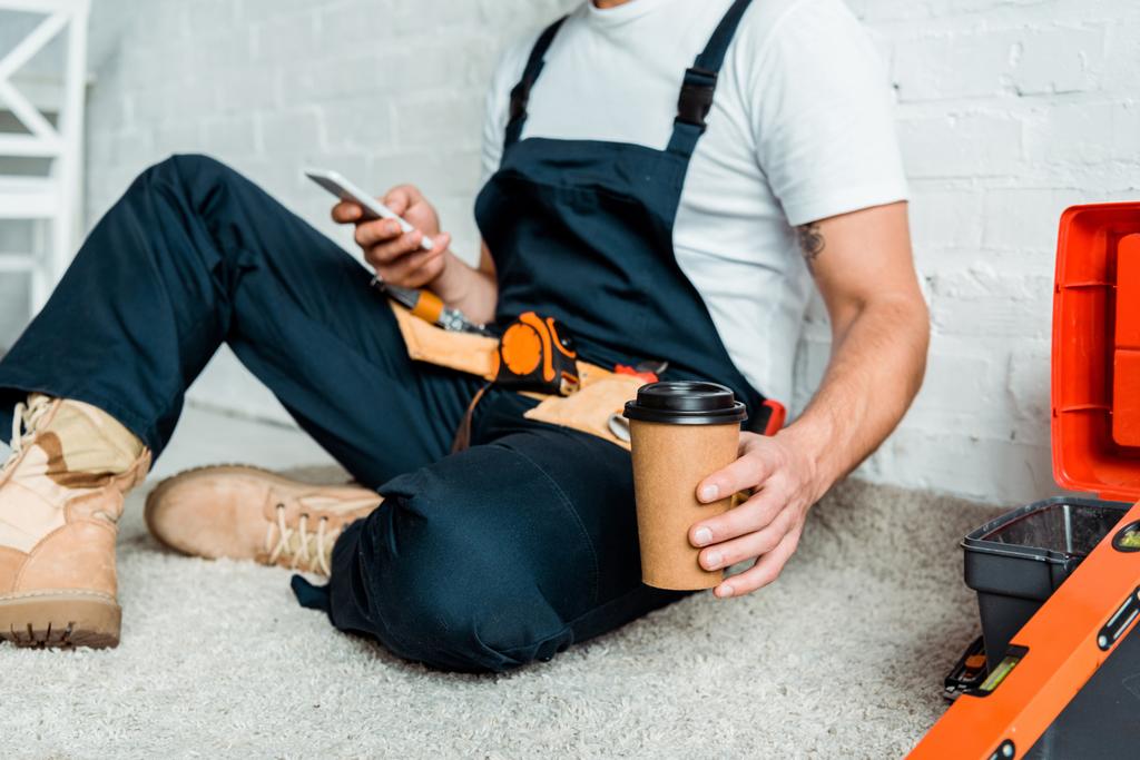 обрезанный вид монтажника, сидящего на ковре, держа бумажную чашку и используя смартфон
  - Фото, изображение