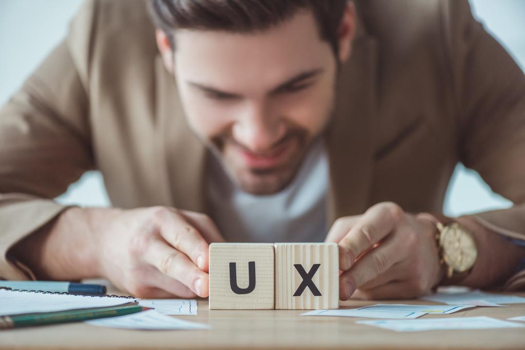 Selective focus of smiling creative designer holding cubes with ux letters at table with layouts - Photo, Image