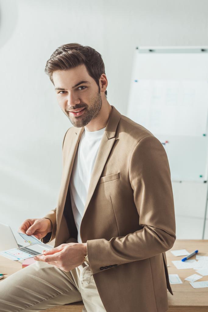 Handsome designer looking at camera while holding sketches by table  - Photo, Image