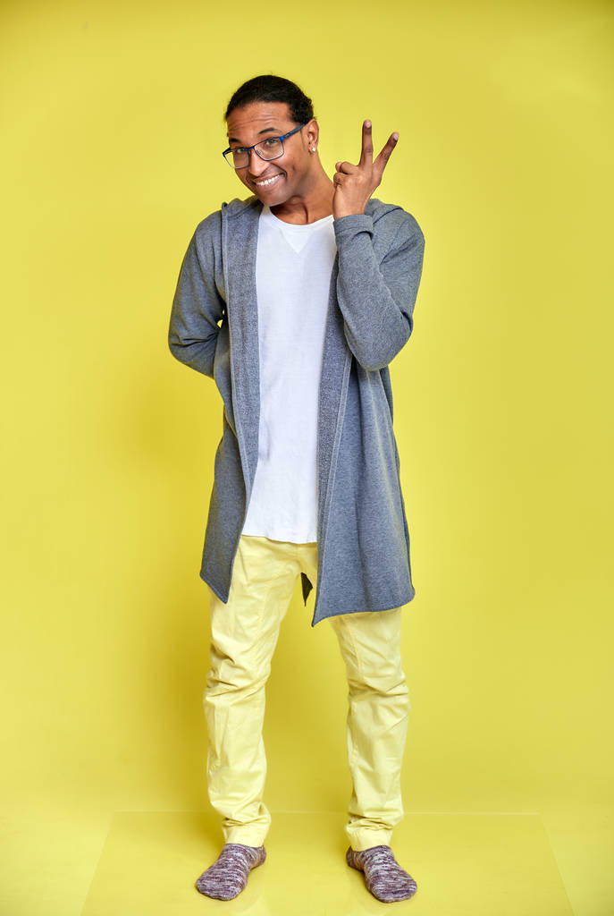 Verticaal portret van een vrolijke jongeman met een bril Afro-Amerikaans met een kort kapsel in gewone kleren op een gele achtergrond in volle lengte. Vlak voor de camera staan - Foto, afbeelding