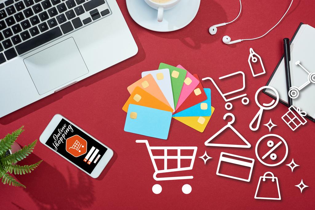 top view of multicolored empty credit cards on red background with online shopping illustration on smartphone near laptop, earphones and coffee - Photo, Image