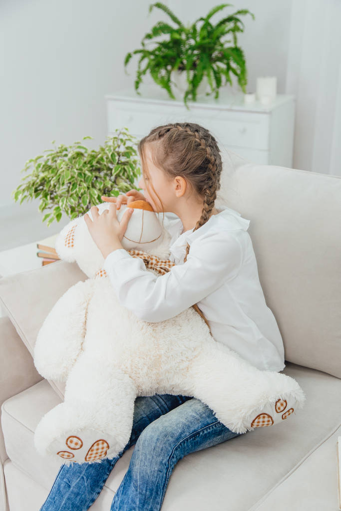 Ευτυχισμένο κοριτσάκι που κάθεται στον καναπέ, κρατώντας ένα μεγάλο αρκουδάκι στα γόνατά της. - Φωτογραφία, εικόνα