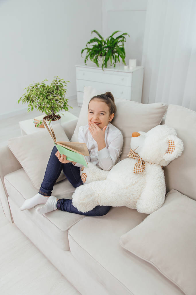 Menina bonito sentado no sofá com ursinho de pelúcia perto dela, lendo conto de fadas, encontrar algo engraçado no livro, rindo alegremente
. - Foto, Imagem