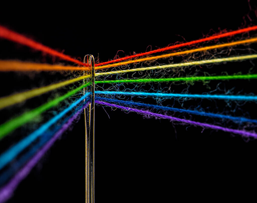 Multicolored threads, forming the colors of the rainbow, are threaded through the eye of a sewing needle, macro - Photo, Image