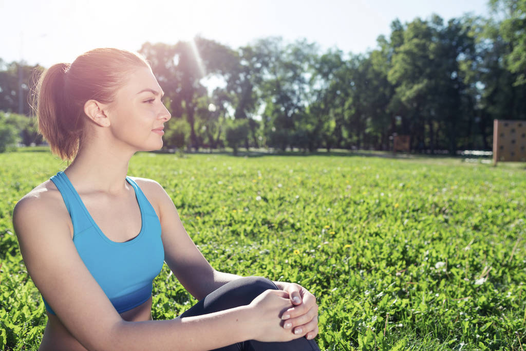 Hermosa chica sonriente en ropa deportiva relajarse en el parque. Mujer joven con sentado en la hierba verde después del entrenamiento. Meditación al aire libre en el soleado día de verano. Ejercicios matutinos y estilo de vida saludable. - Foto, imagen