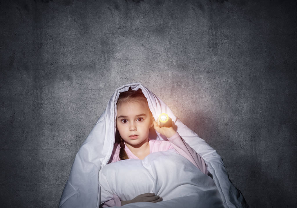Налякана дівчина з ліхтариком ховається під ковдрою. Налякана дитина лежить у ліжку вдома. Нічні жахи від дитини. Маленька дівчинка бояться темряви. Дитина в піжамі на тлі сірої стіни
. - Фото, зображення