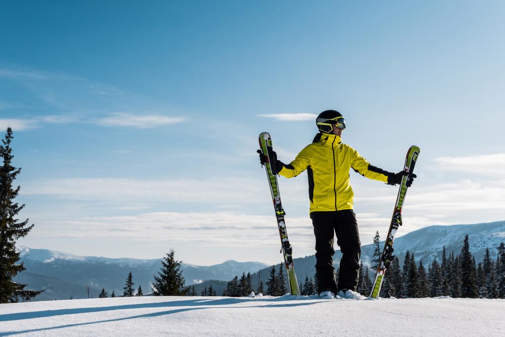 σκιέρ κρατώντας σκι και στέκεται ενάντια στον ουρανό στα βουνά - Φωτογραφία, εικόνα