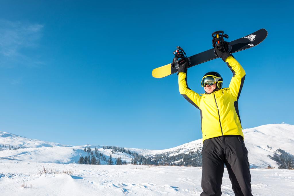χαρούμενος αθλητής στο κράνος κρατώντας snowboard πάνω από το κεφάλι - Φωτογραφία, εικόνα